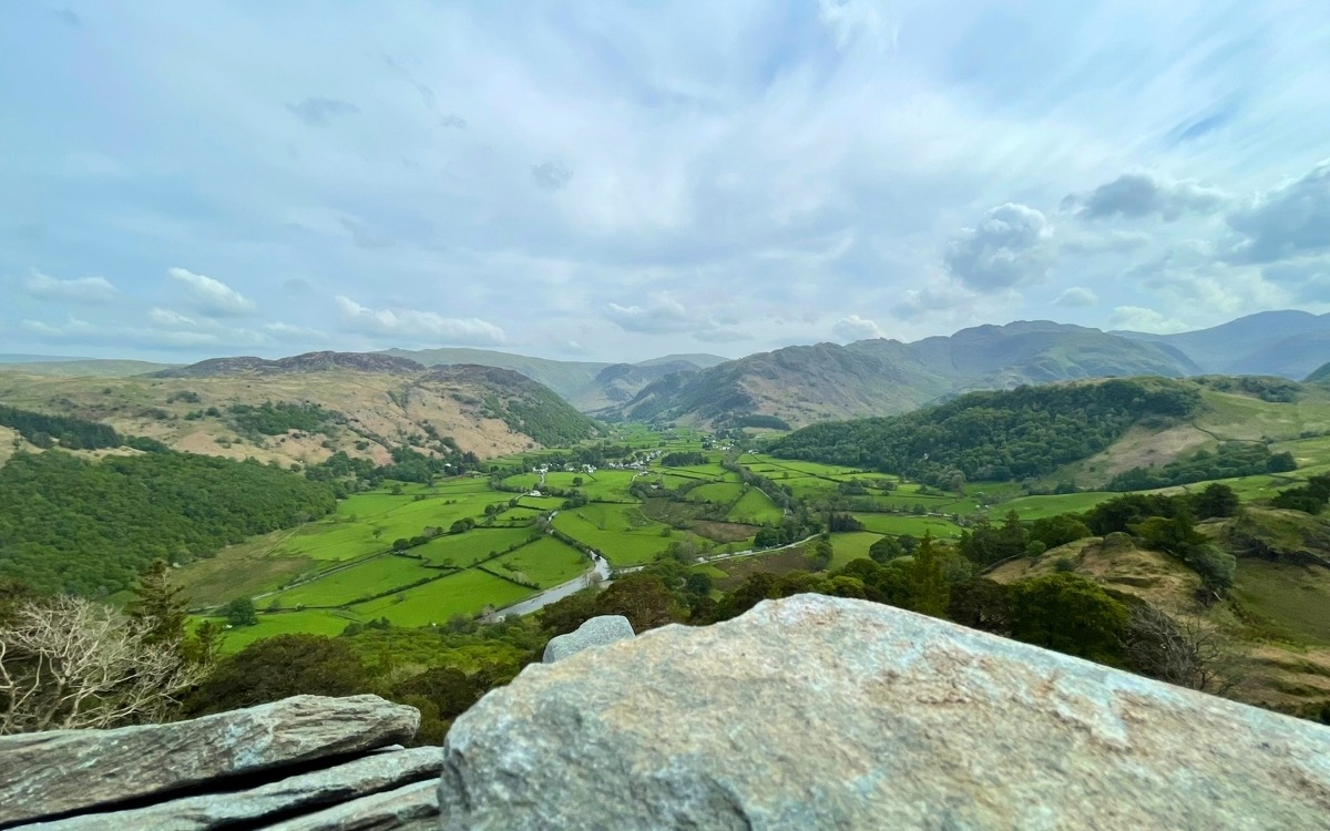 Elevated panoramic view over rocky slate crags looking south across the patchwork green fields and winding river of the Borrowdale valley towards Rosthwaite and the distant high fells of Seathwaite.