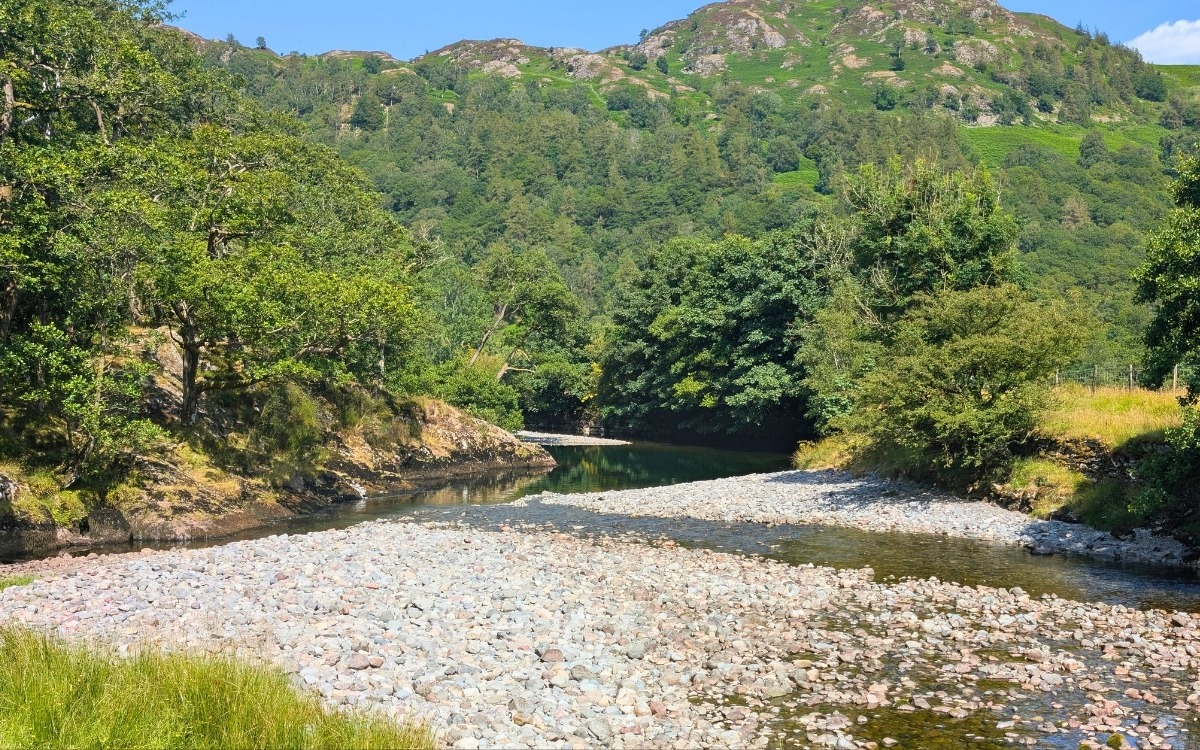 Wide pebble and gravel shingle bank along the River Derwent with clear emerald water, framed by lush green trees and sunlit fells on the wild riverside path to Grange.