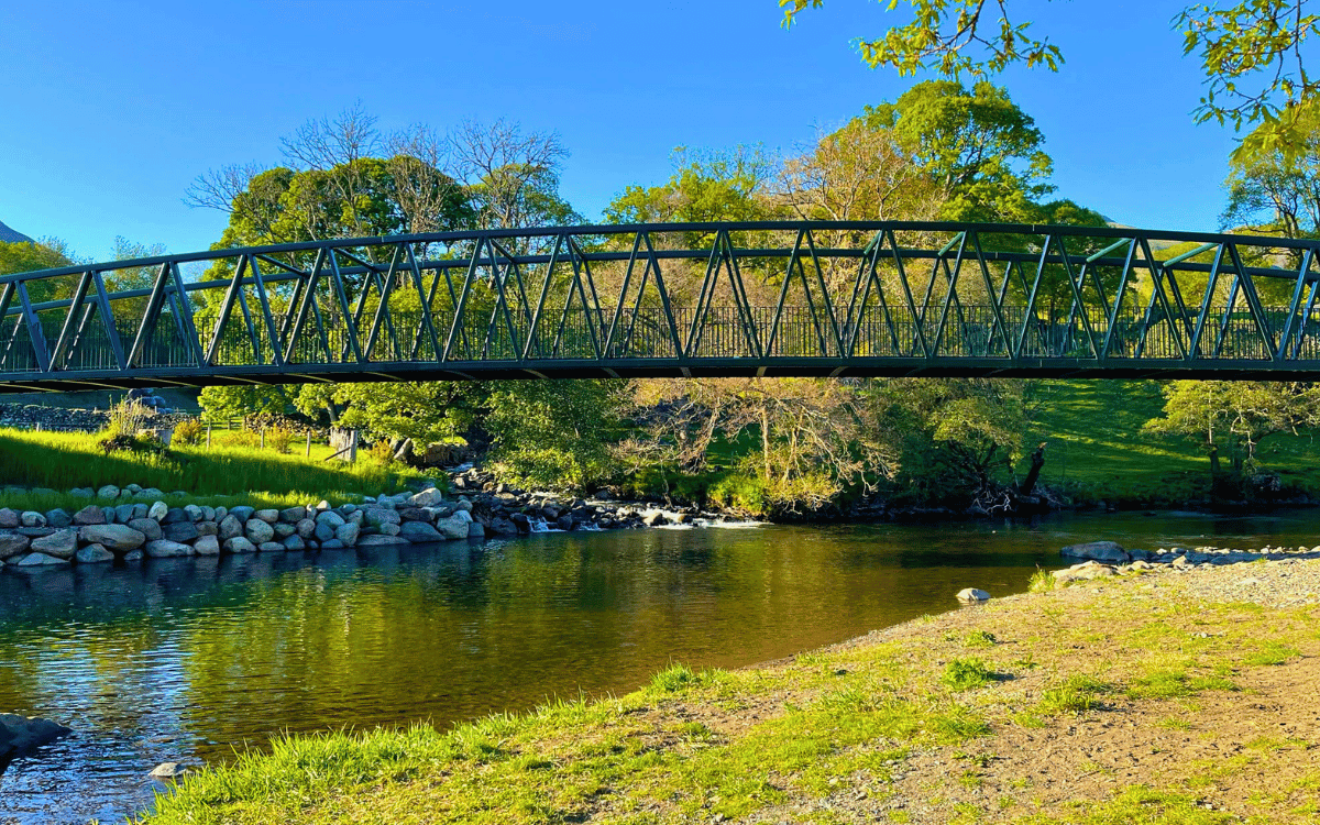 A peaceful view from a grassy and sandy shoreline looking across the calm, clear waters of the River Greta. A modern, dark green metal bridge from the Keswick to Threlkeld trail spans the river, framed by bright green summer trees under a clear blue sky.
