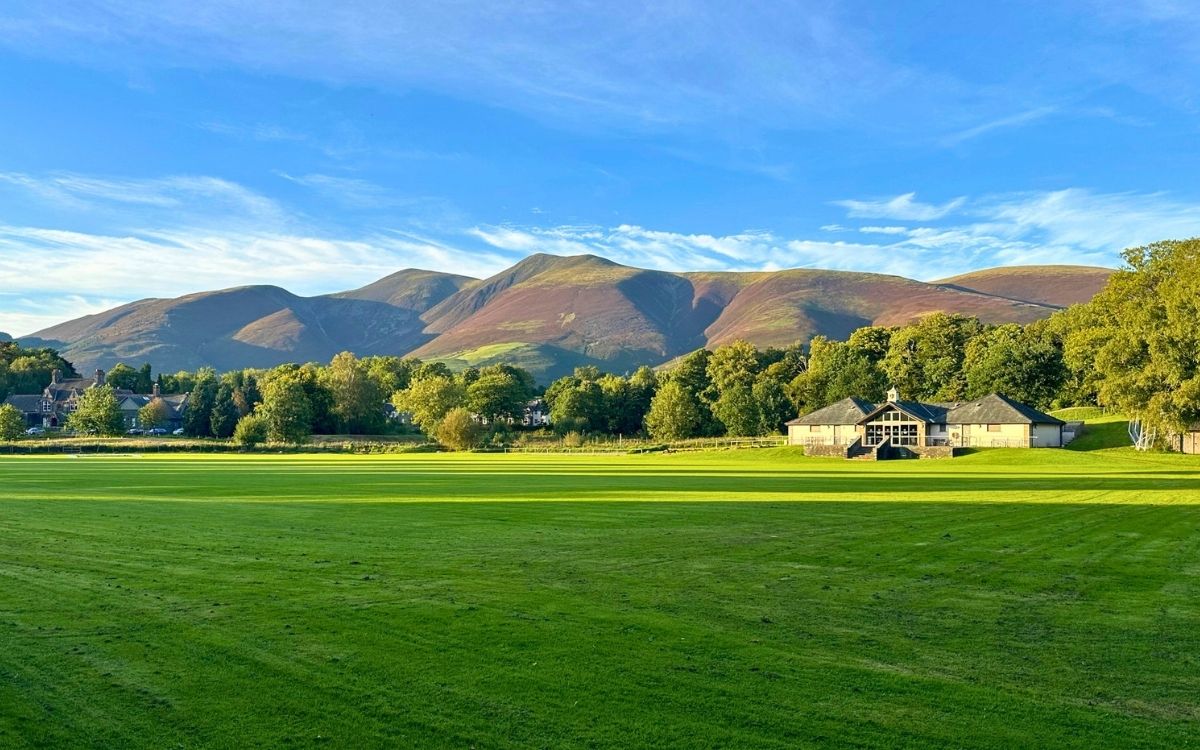 Panoramic view of Skiddaw and the Northern Fells rising behind Keswick across the manicured green grounds of Fitz Park under a blue sky in Keswick.