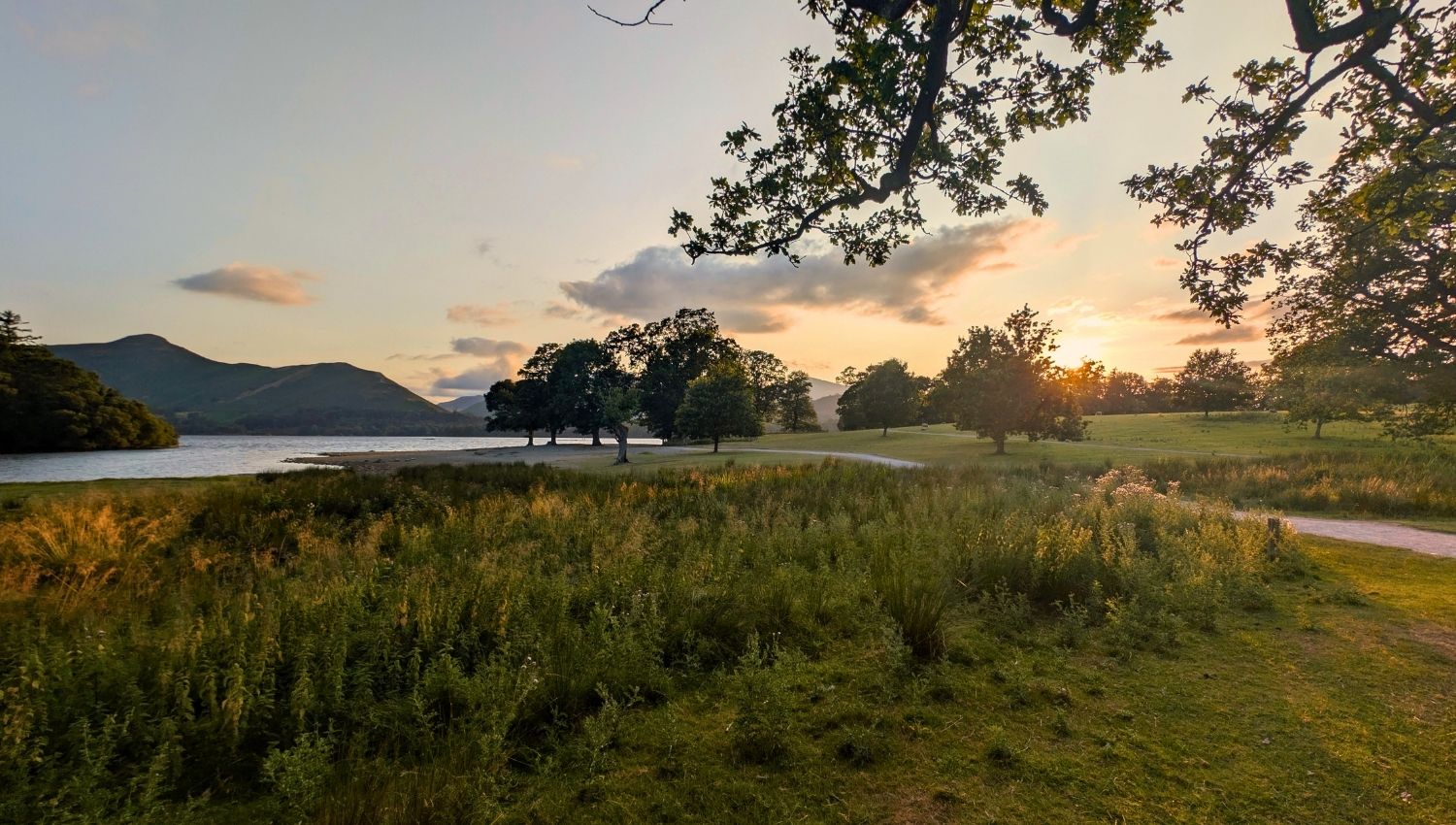 A warm summer sunset casting an orange glow over Strandshag Bay on the pebbled shores of Derwent Water, with the silhouette of Catbells mountain in the background.