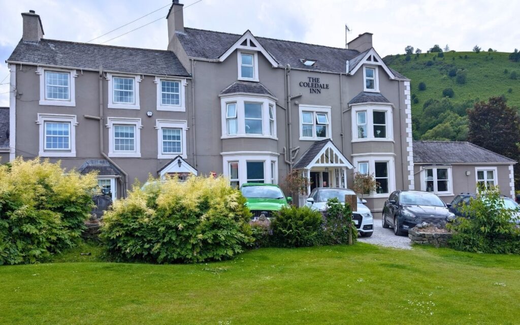 View of the historic Coledale Inn coaching inn from a wooden picnic bench in the pub garden, showing patrons sitting outside with the green Cumbrian fells rising in the background in Braithwaite near Keswick.