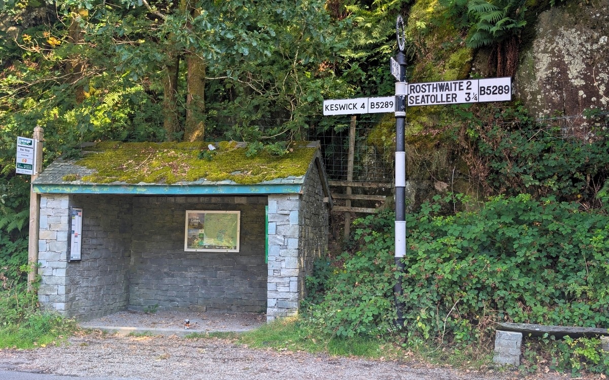 Traditional Lakeland slate stone bus shelter with a moss-covered roof and a black-and-white fingerpost road sign pointing towards Keswick, Rosthwaite, and Seatoller in Grange in Borrowdale.