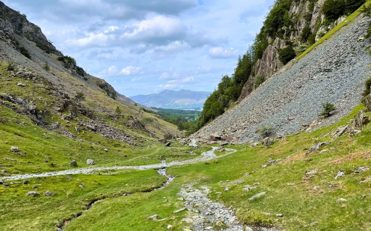 Gravel walking track cutting through a mountain pass between steep slate scree slopes, looking down towards Derwentwater with the distant peak of Skiddaw under a blue summer sky.