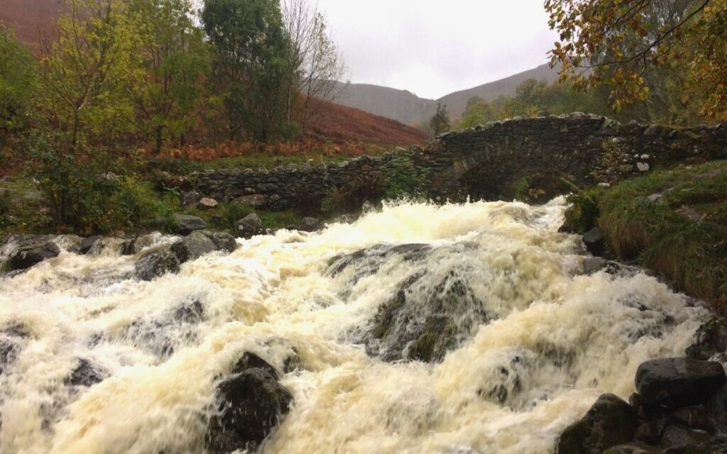 Historic stone packhorse arch of Ashness Bridge framed by a roaring, powerful white-water cascade surging over boulders in Borrowdale.