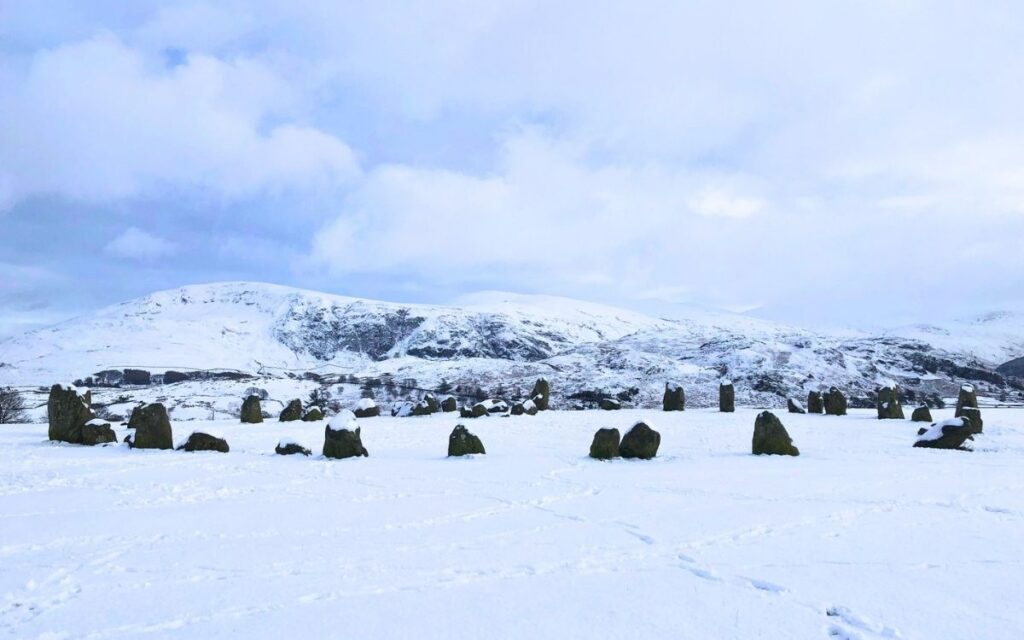 Ancient megalith standing stones of Castlerigg Stone Circle dusted in deep white snow with snow-covered Lake District fells in the background.