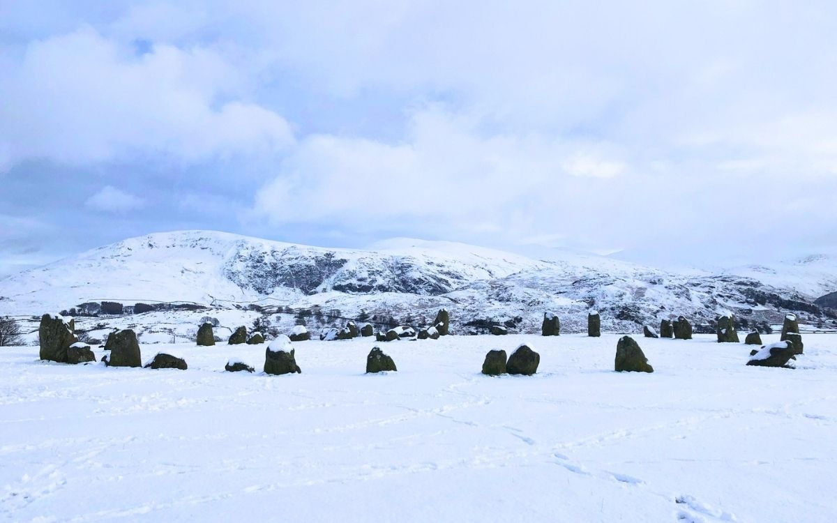Ancient megalith standing stones of Castlerigg Stone Circle dusted in deep white snow with snow-covered Lake District fells in the background.