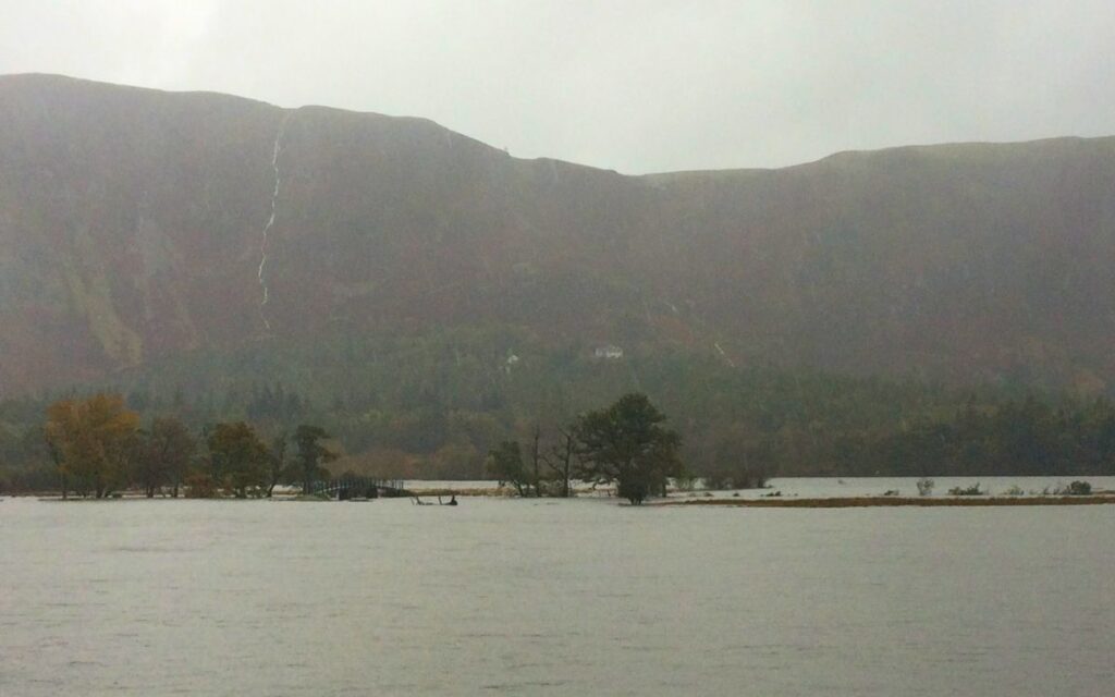 Expansive grey flood water at the head of Derwent Water with the Chinese Bridge completely submerged beneath the lake surface under misty fells.