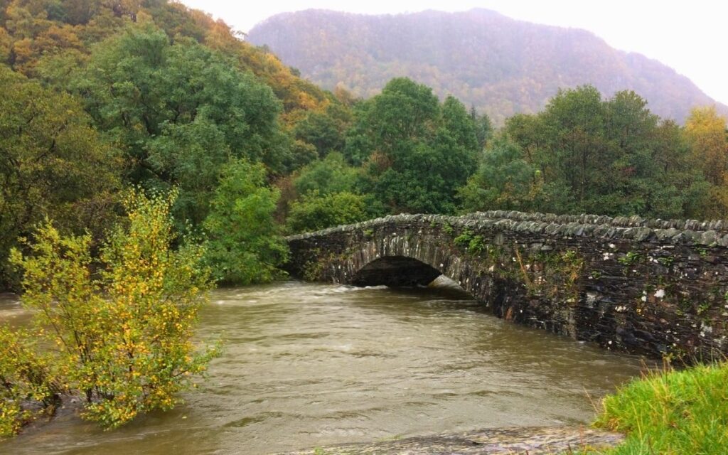 Double-arch stone Grange Bridge in Borrowdale spanning a churning, swollen brown River Derwent beneath mist-covered valley fells.