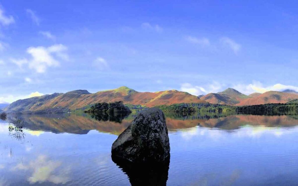 Large mossy boulder emerging from still, high flood waters of Derwent Water with Catbells and the western fells reflected on the lake surface.